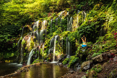 Rear view of woman practicing yoga while sitting on rock against waterfall in forest