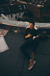 High angle full length of thoughtful man using mobile phone while relaxing on terrace during party at dusk