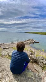 Rear view of woman sitting on rock by sea against sky