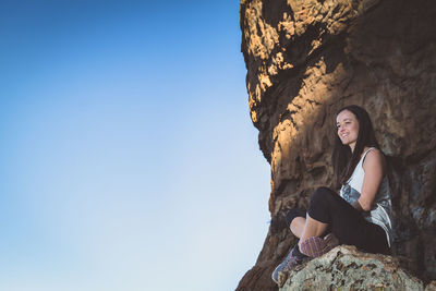 Low angle view of smiling woman sitting on rock against clear blue sky
