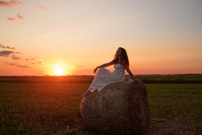 Woman sitting on haystack against sky during sunset