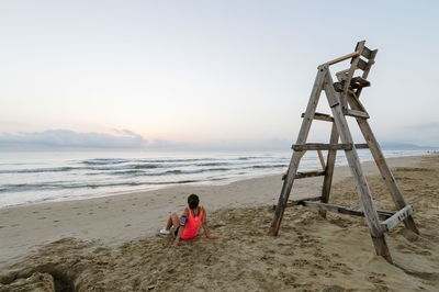 Rear view of woman sitting by wooden ladder at beach against sky