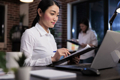 Young businesswoman holding file folder looking at laptop screen in office