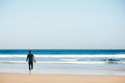 Rear view of man with surfboard walking at beach against clear sky