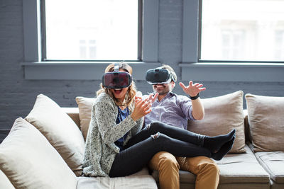 Happy coworkers wearing virtual reality simulators while sitting on sofa in office