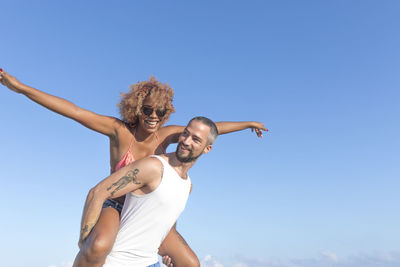 Low angle view of man piggybacking cheerful girlfriend against blue sky