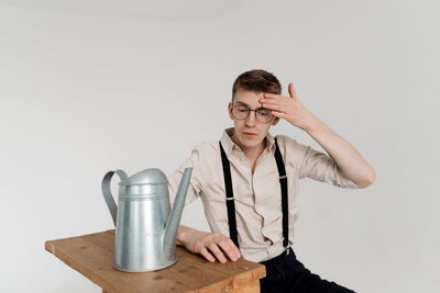 Young man with watering can sitting on ladder against white background