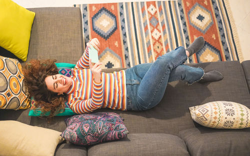 High angle view of cheerful woman taking selfie while lying on sofa at home