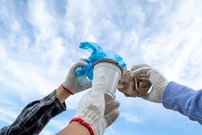 Low angle view of people wearing glove holding garbage against sky