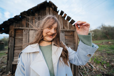 Beautiful girl with long hair in a grey trench coat next to an old wooden house outdoors in spring