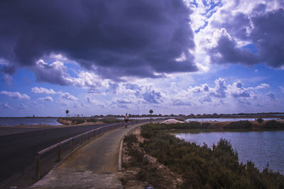 Rear view of man riding bicycle on road leading river against cloudy sky