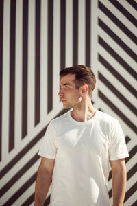 Serious sportsman in activewear standing on street in summer and looking away against stripe wall background