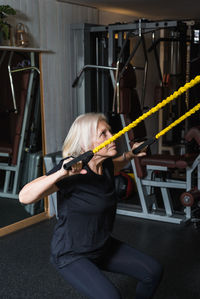 Side view of elderly female athlete in sportswear with gray hair working out with straps in gym