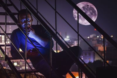 Portrait of young man using phone while sitting on steps against illuminated city at night