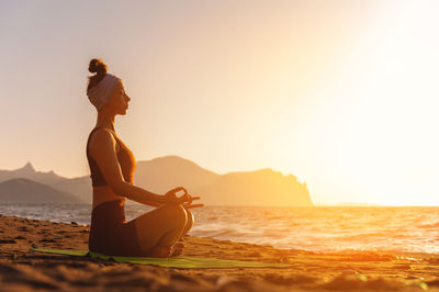 Rear view of woman sitting on beach against sky during sunset