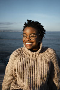 Smiling woman wearing eyeglasses looking away while sitting against sea