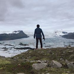 Rear view of hiker with camera standing on field by glaciers against cloudy sky