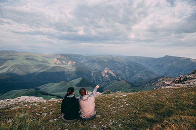 Two guys are sitting and looking into the distance and the mountains, discussing something and showing somewhere far into the distance with his hand.