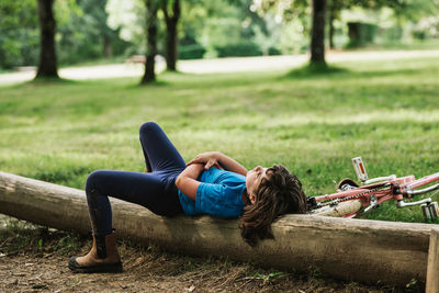 Side view of relaxed thoughtful girl in casual clothes lying on log near bike while spending sunny summer day in park