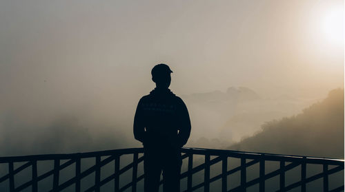 Rear view of silhouette man standing by railing against sky