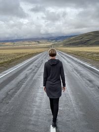 A young woman in a long hooded sweatshirt walking away into the distance on a cloudy fall day. 