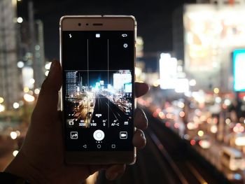 Cropped hand photographing illuminated cityscape through mobile phone at night