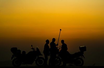Silhouette friends with monopod standing by motorcycles at sunset