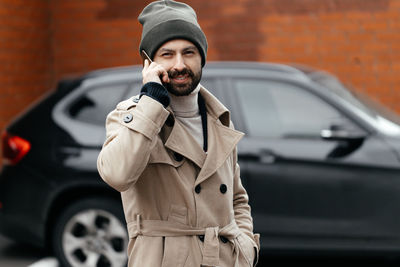 Close-up of a young, smiling, happy, laughing hipster with a beard in a raincoat and hat, 