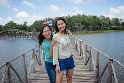 Portrait of smiling friends standing on footbridge over lake