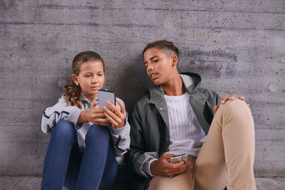 Sister showing her mobile phone to brother while sitting against wall