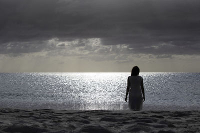 Rear view of woman standing at beach against sky during sunset