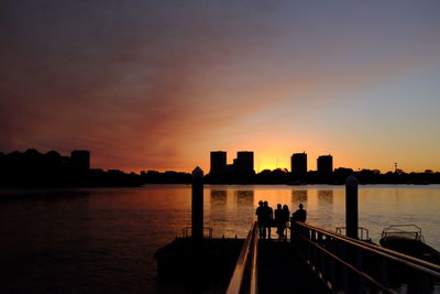 Silhouette people on sea by city against sky during sunset