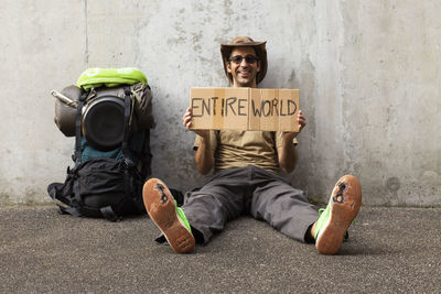 Portrait of man holding cardboard with entire world text while sitting against wall 