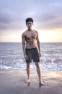 Portrait of shirtless man standing at beach against sky