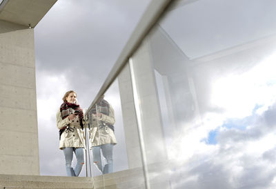 Low angle view of woman holding mobile phone while standing by railing against sky
