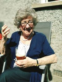 Portrait of smiling woman holding drink while sitting on chair during sunny day