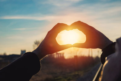 Cropped hands of man making heart shape against sky during sunset