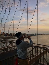 Rear view of man photographing east river while standing on brooklyn bridge during sunset