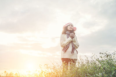 Thoughtful woman in warm clothing standing on field against cloudy sky