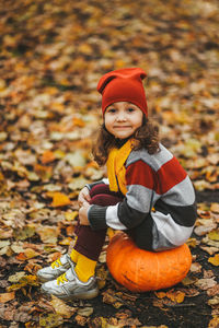Happy cute little girl child in warm clothes sitting on a pumpkin in the autumn forest outdoors