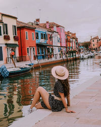 Rear view of woman sitting by canal against buildings