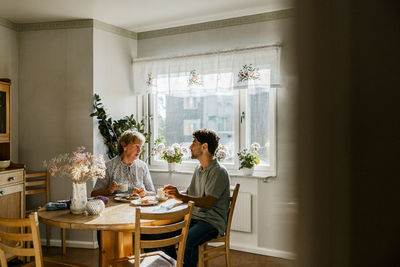 Young male nurse having breakfast with senior woman while sitting at dining table in kitchen