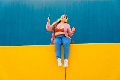 Full length of woman with arms raised sitting on yellow wall