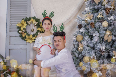 Portrait of young man with pregnant girlfriend sitting by christmas tree at home