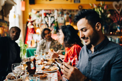 Smiling young man text messaging on smart phone while sitting with friends at table in restaurant during dinner party