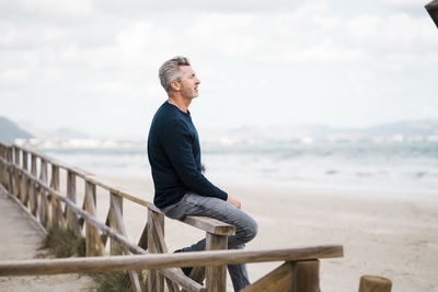 Thoughtful mature man sitting on railing at beach