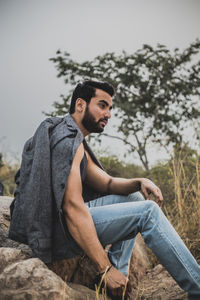 Young man sitting on tree against sky