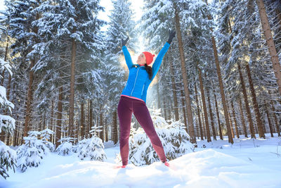 Mid adult woman with arms raised standing on snow covered field against trees in forest
