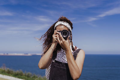 Portrait of young woman photographing sea against sky