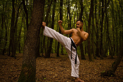 Shirtless man practicing karate against trees in forest